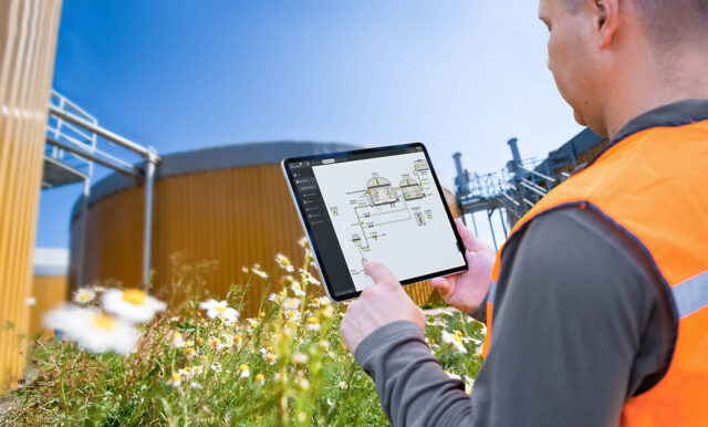 Osoba trzyma tablet przed instalacją przemysłową na zewnątrz.